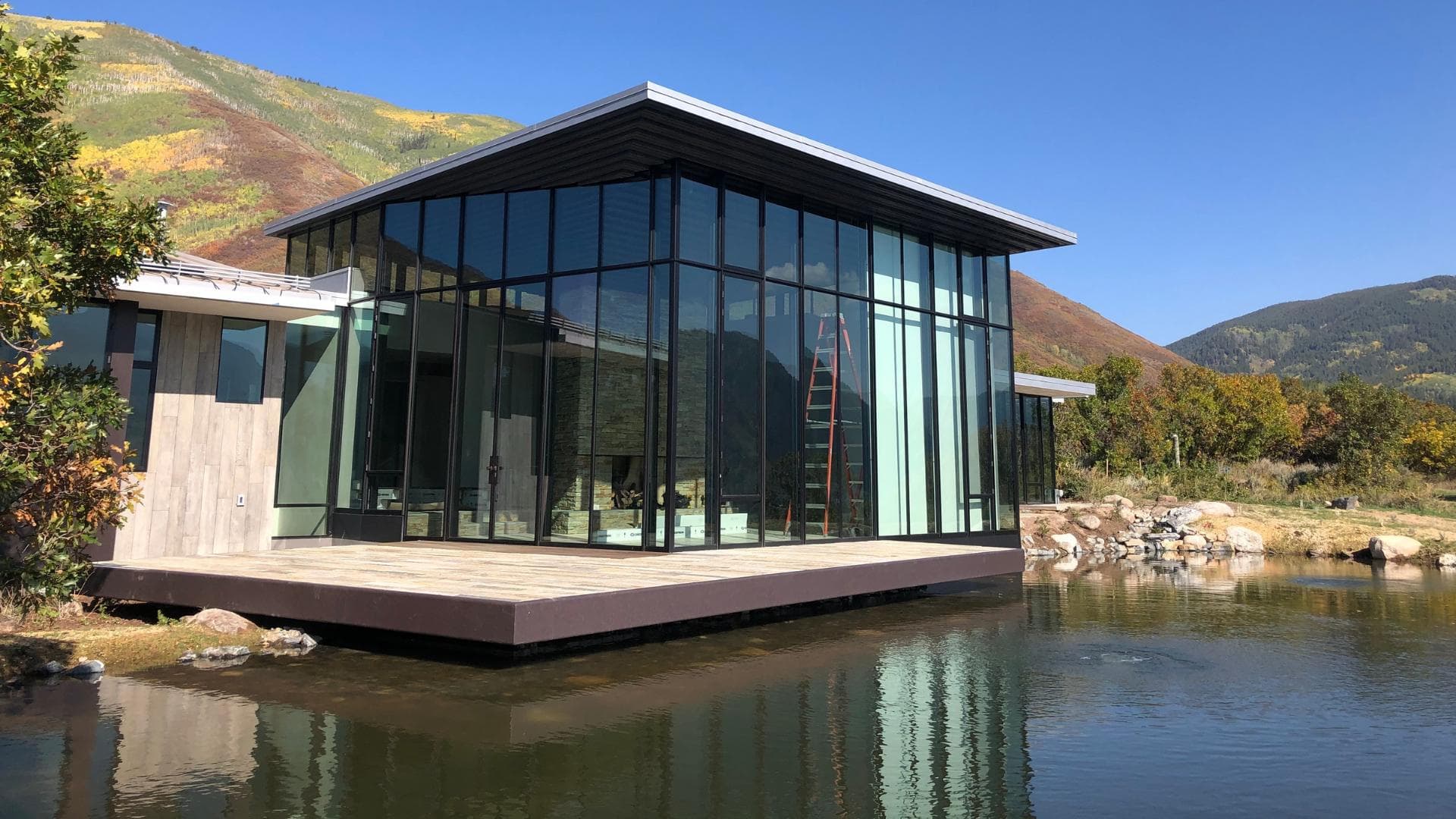 A glass pavilion set over a pond, with fall color on the ridge behind